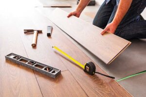 Person vacuuming a carpeted living room beside a small entry mat and cleaning supplies.